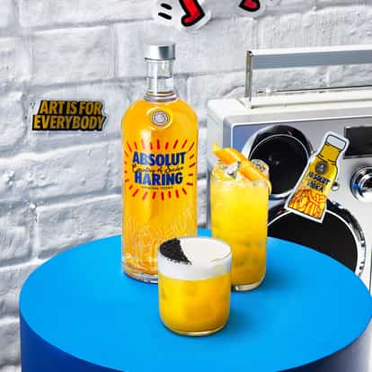 Absolut Haring Fizz and an Absolut Haring Collins sitting on a round blue table next to a bottle of Absolut Haring and a sticker on the wall saying 'Art is for everybody'-