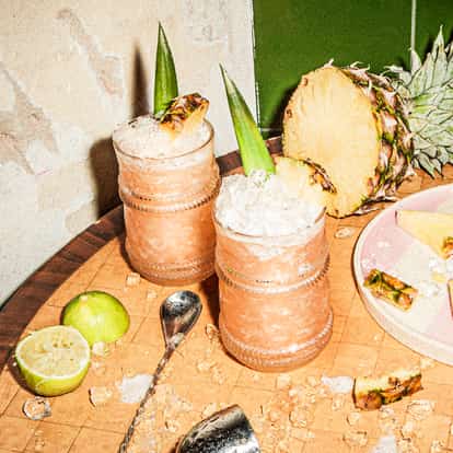 Jungle Bird cocktails surrounded by fresh pineapple, lime halves and a bar spoon.
