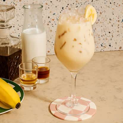 Iced banana cocktail in a stemmed glass garnished with a banana slice, with milk, liquor, syrup, and a fresh banana displayed beside it.