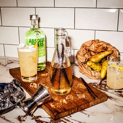 Vanilla Syrup in a glass bottle with vanilla pods on a wooden board beside a jigger and an Absolut Pears bottle